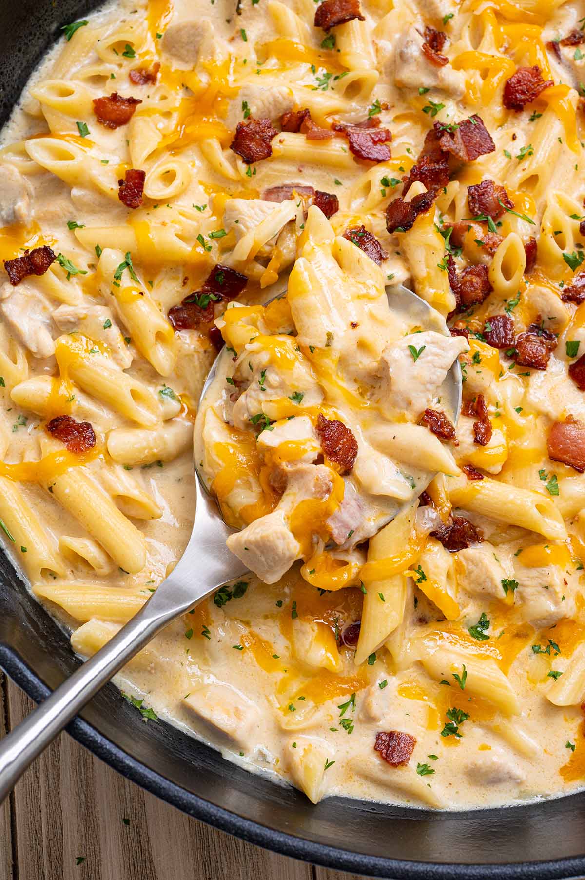Close-up of a spoon lifting crack chicken penne with melted cheddar and crispy bacon from a cast iron skillet.