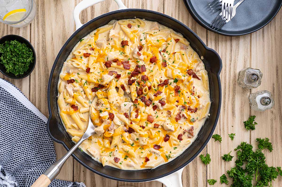 Crack chicken penne in a white cast iron skillet topped with cheddar cheese, crispy bacon and fresh parsley.