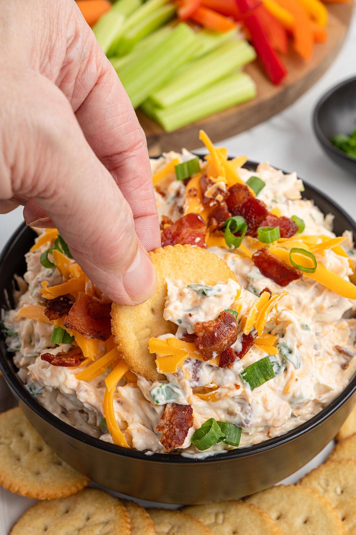 Hand dipping a Ritz cracker into million dollar dip topped with cheddar cheese, bacon, and green onions.