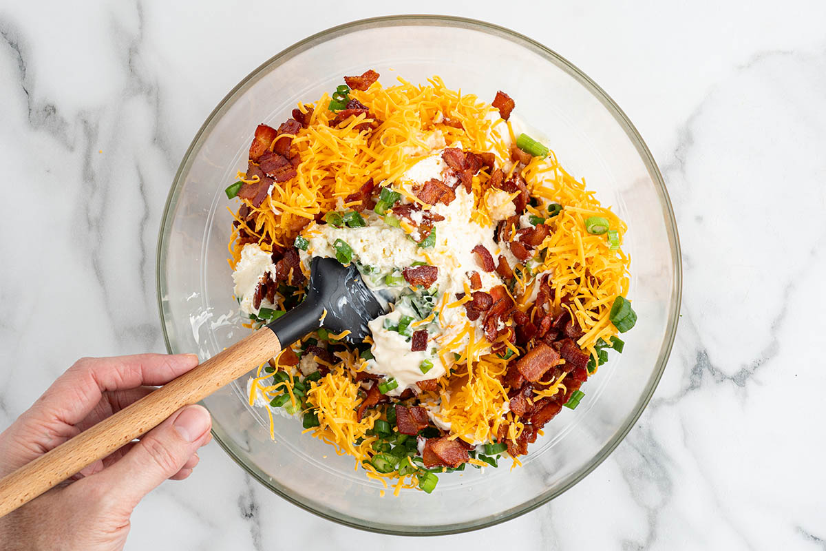 Hand stirring million dollar dip ingredients together in a glass bowl with a rubber spatula.