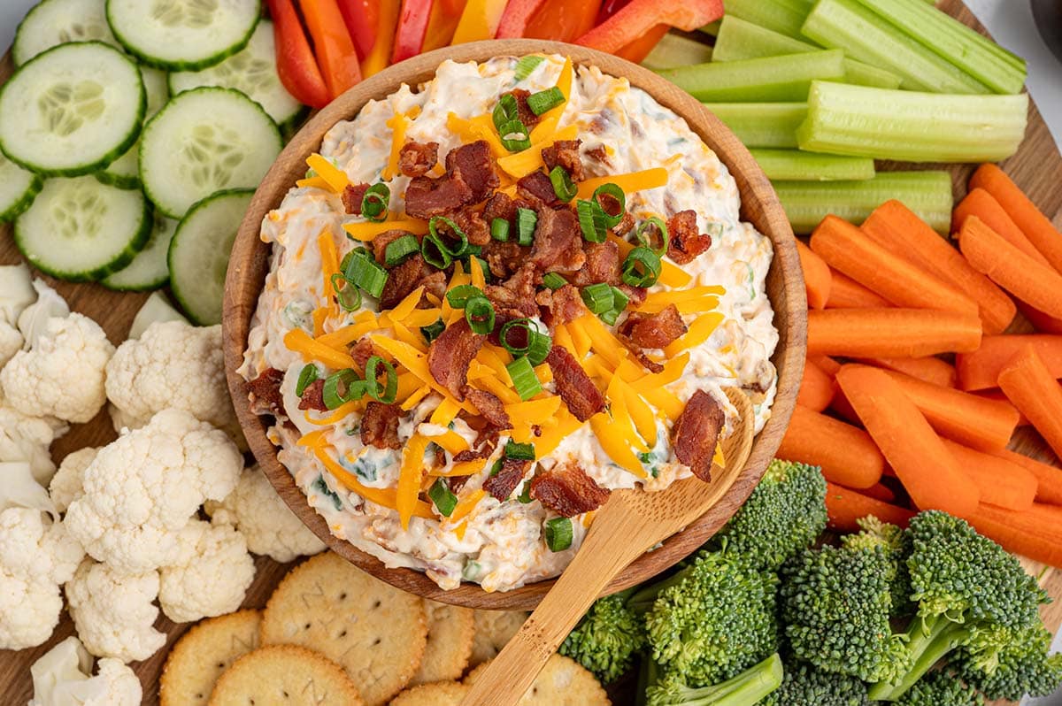 Overhead view of million dollar dip with a wooden spoon in a wooden bowl, surrounded by fresh vegetables and crackers on a round serving board.