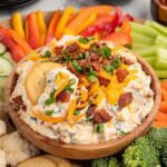 Million dollar dip in a wooden bowl topped with cheddar cheese, bacon, and green onions, surrounded by crackers, fresh vegetables, and dipping accompaniments.