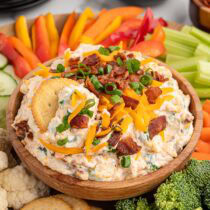 Million dollar dip in a wooden bowl topped with cheddar cheese, bacon, and green onions, surrounded by crackers, fresh vegetables, and dipping accompaniments.