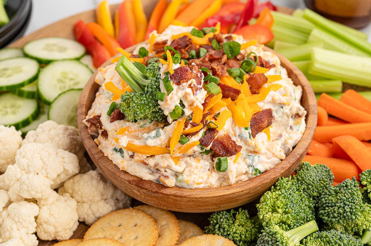 Million dollar dip loaded with cheddar cheese, bacon, and green onions in a wooden bowl, served with fresh vegetables and crackers.