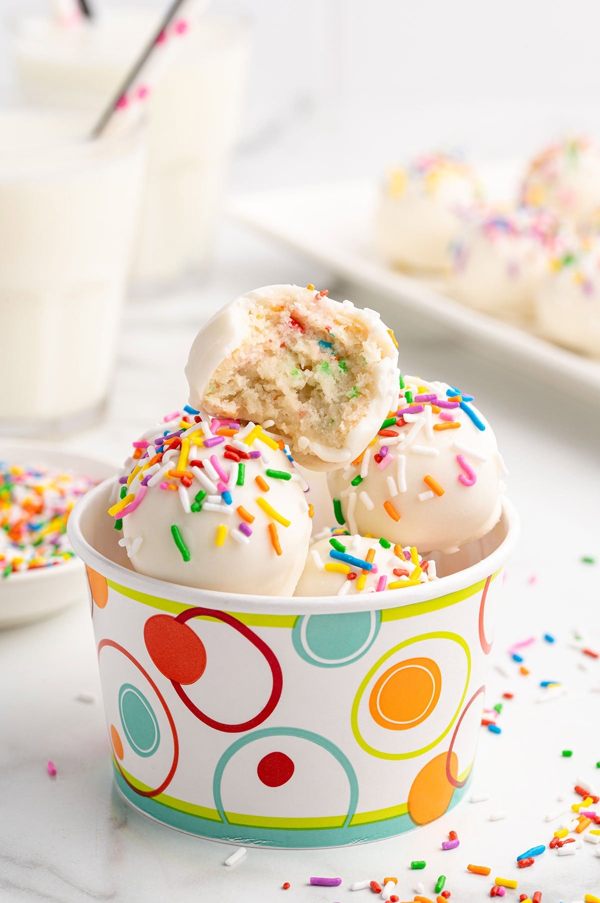 No-bake cheesecake balls dipped in white chocolate and topped with rainbow sprinkles in a colorful cup, with one bitten open to show the funfetti filling.