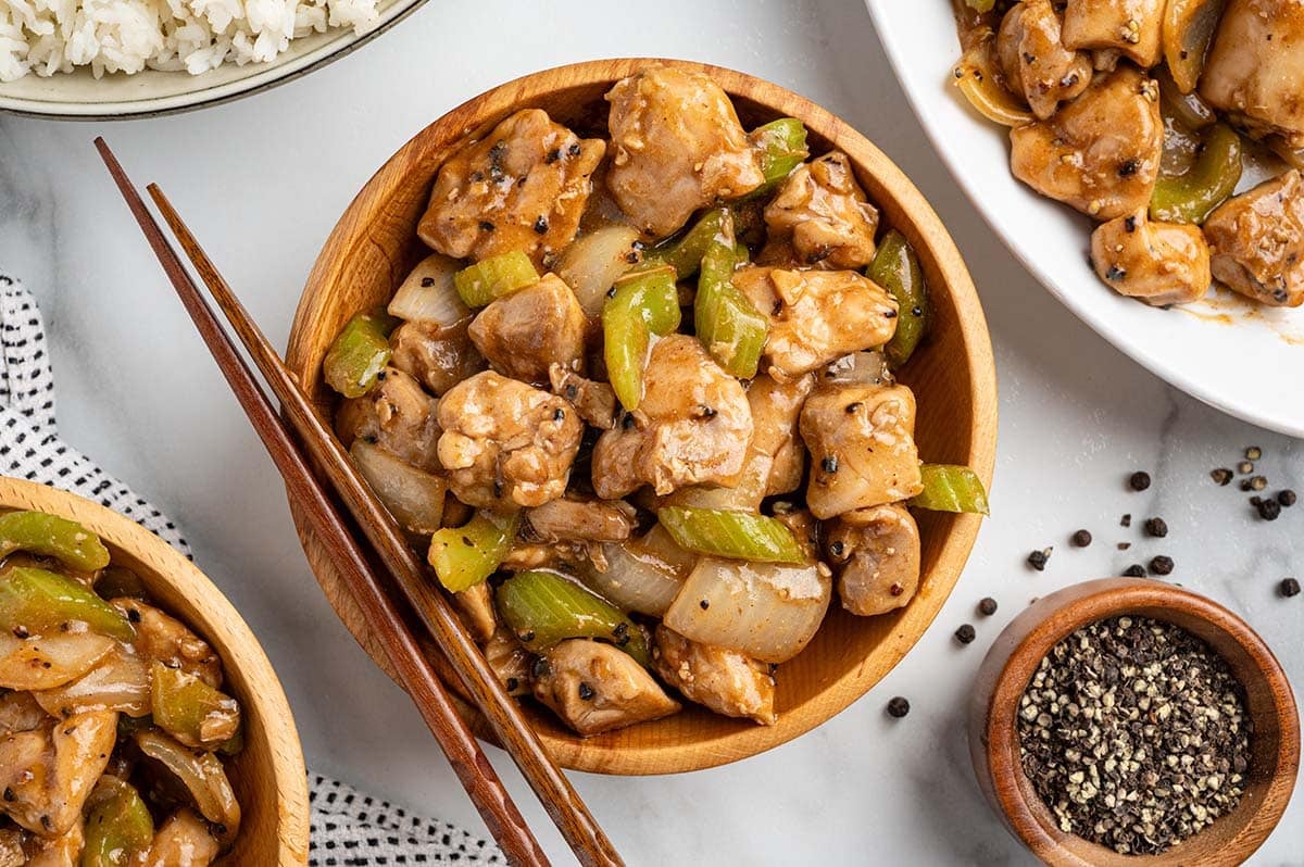 Overhead view of Panda Express pepper chicken in a wooden bowl with chopsticks, surrounded by a bowl of black pepper, steamed rice, and additional servings.