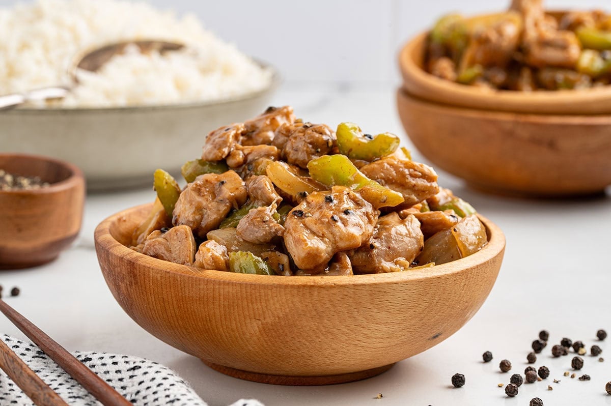 Overhead view of Panda Express pepper chicken in a wooden bowl with chopsticks, surrounded by a bowl of black pepper, steamed rice, and additional servings.11:08 AMCopycat Panda Express pepper chicken in a wooden bowl with celery and onions, with black peppercorns, steamed rice, and chopsticks nearby.