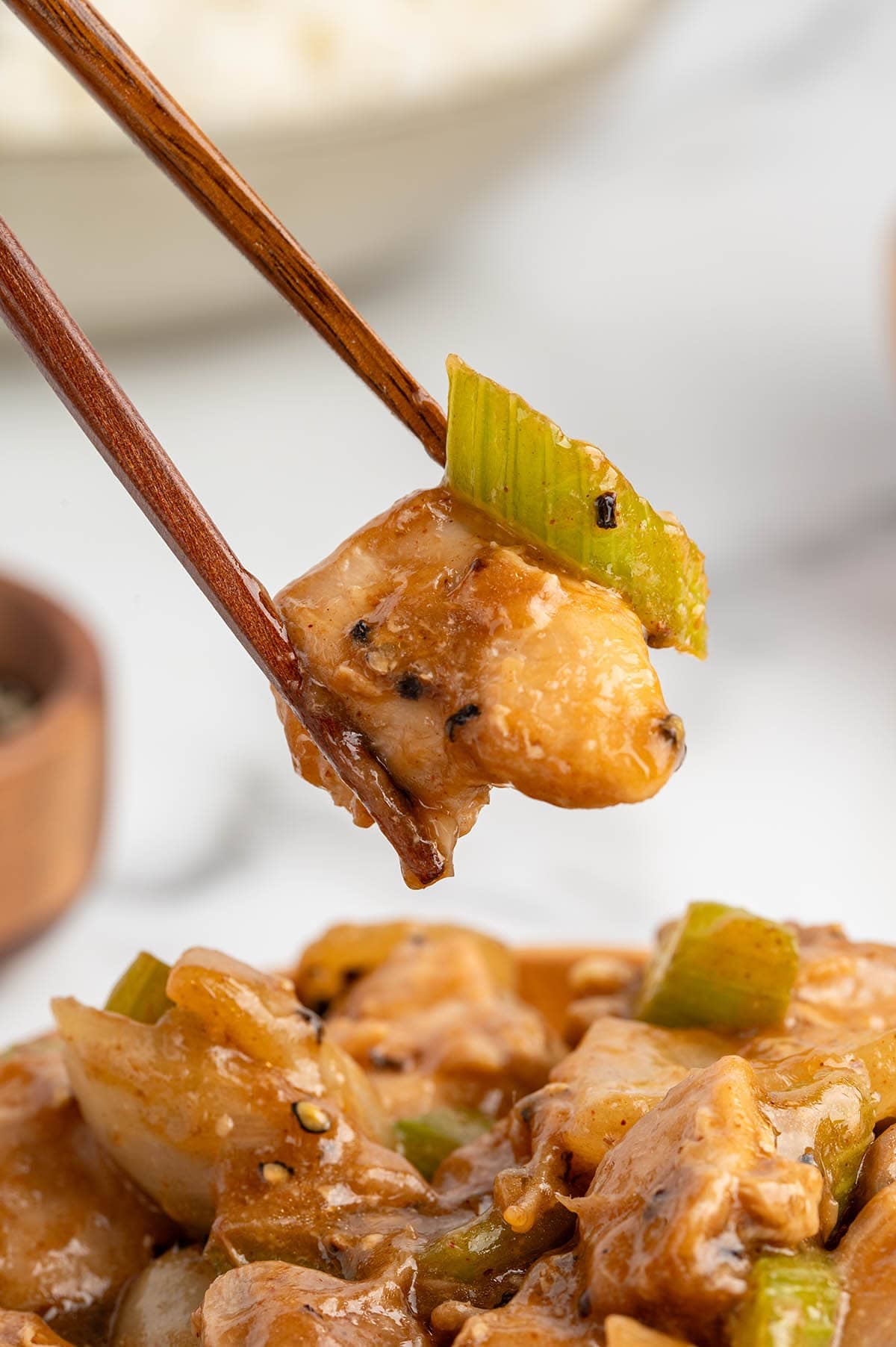 Close-up of chopsticks holding a piece of pepper chicken and celery coated in black pepper sauce.