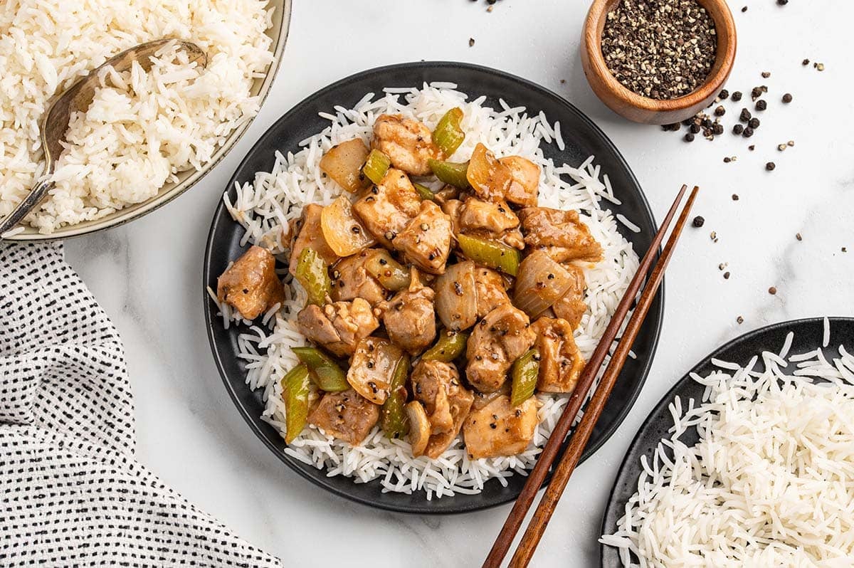 Copycat Panda Express pepper chicken served over steamed white rice on a black plate with chopsticks, with a wooden bowl of black pepper nearby.