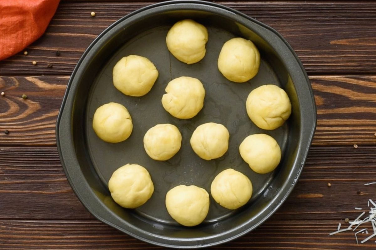 Top-down view of stuffed garlic dinner roll dough balls arranged in a round baking pan before baking.