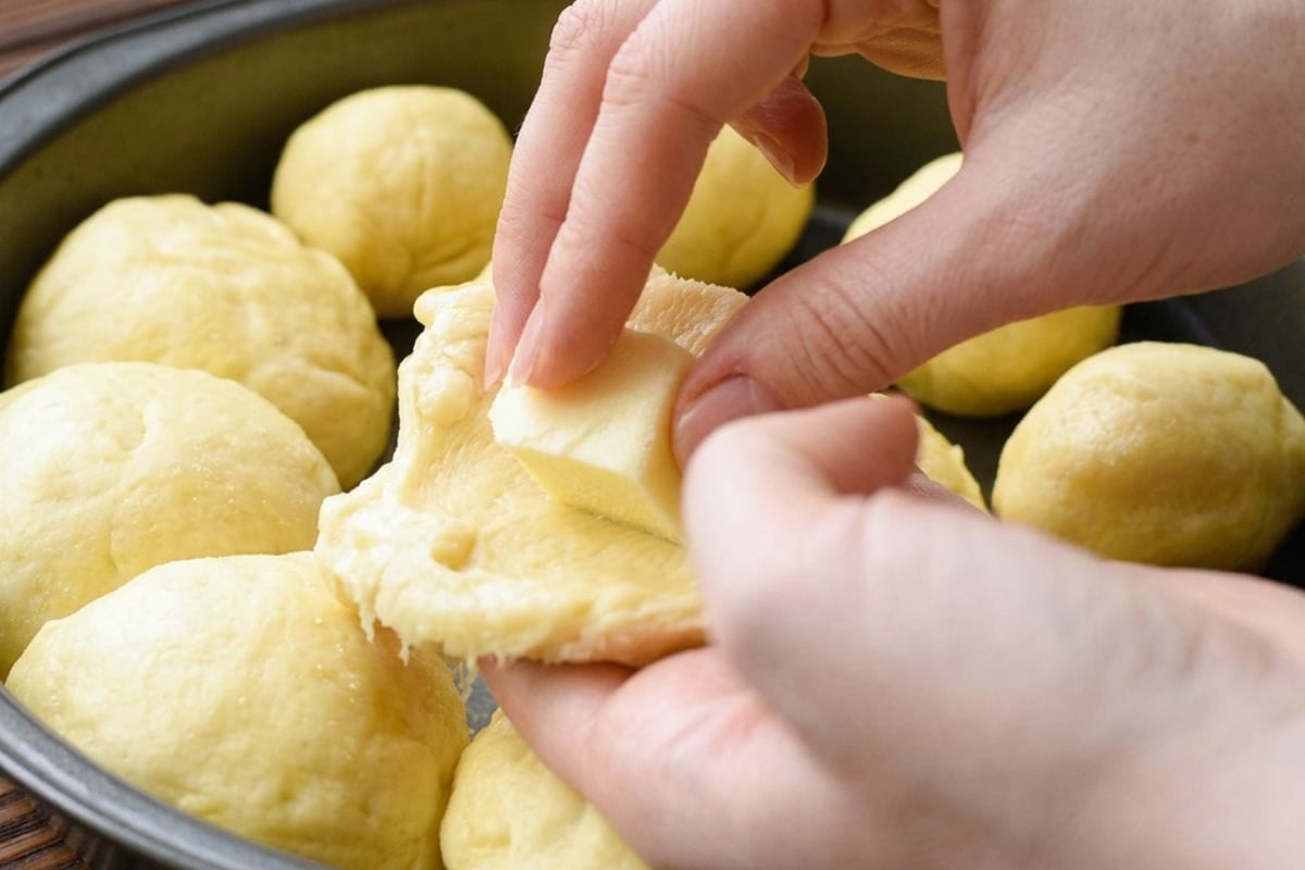 Hands stretching soft dough around a cube of cheese while shaping stuffed garlic dinner rolls in a baking pan.