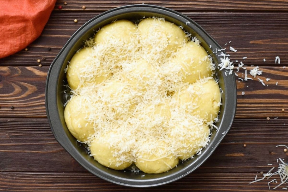 Top-down view of risen garlic dinner rolls in a round pan topped generously with shredded parmesan cheese before baking.