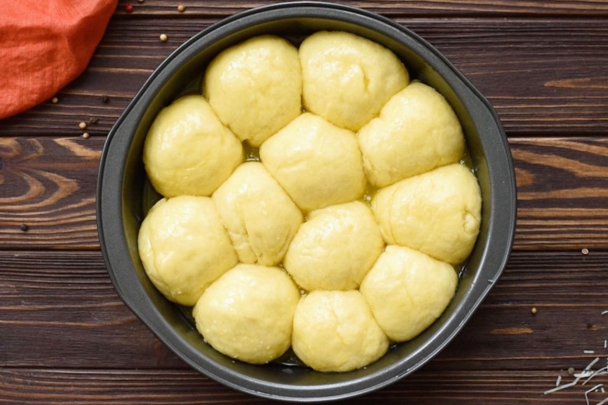 Top-down view of soft risen dinner roll dough in a round baking pan brushed with garlic butter before adding cheese and baking.