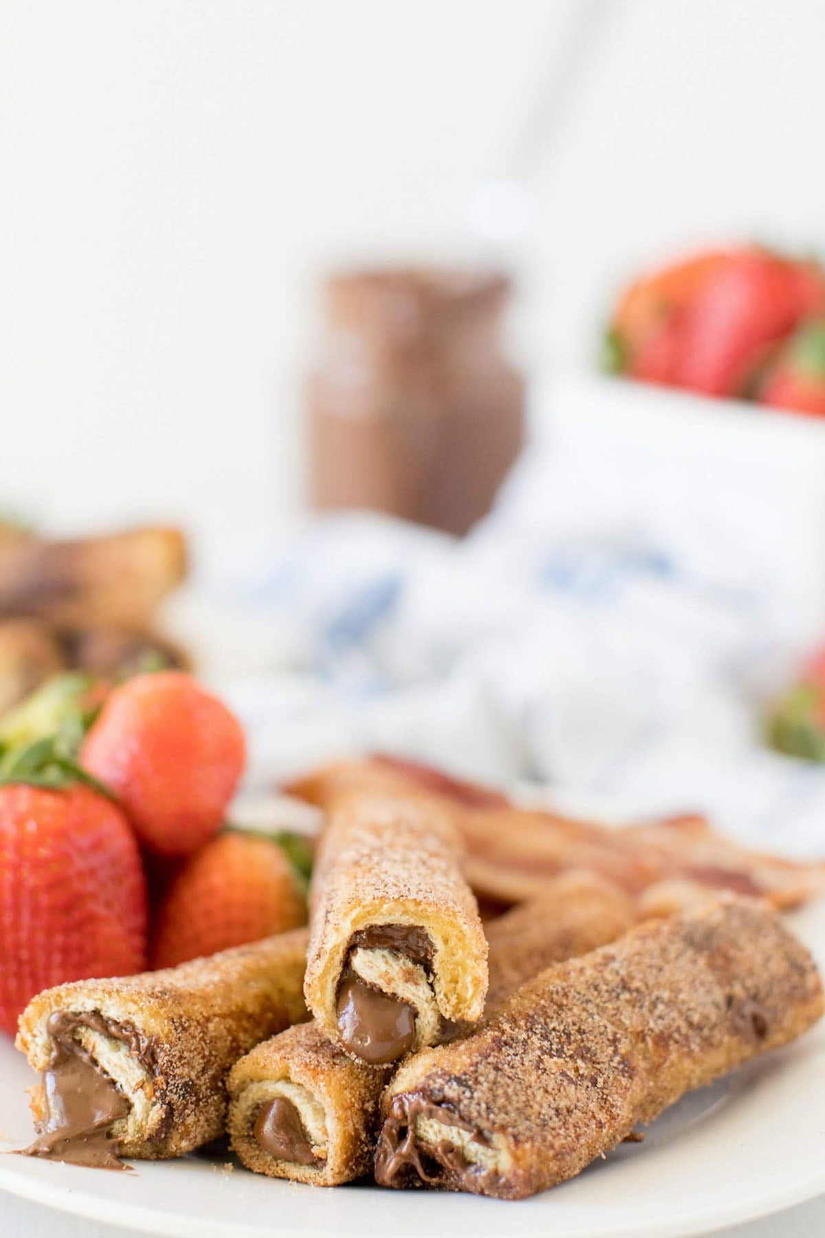 Nutella French toast roll ups coated in cinnamon sugar with melted chocolate hazelnut filling, served with fresh strawberries in the background.
