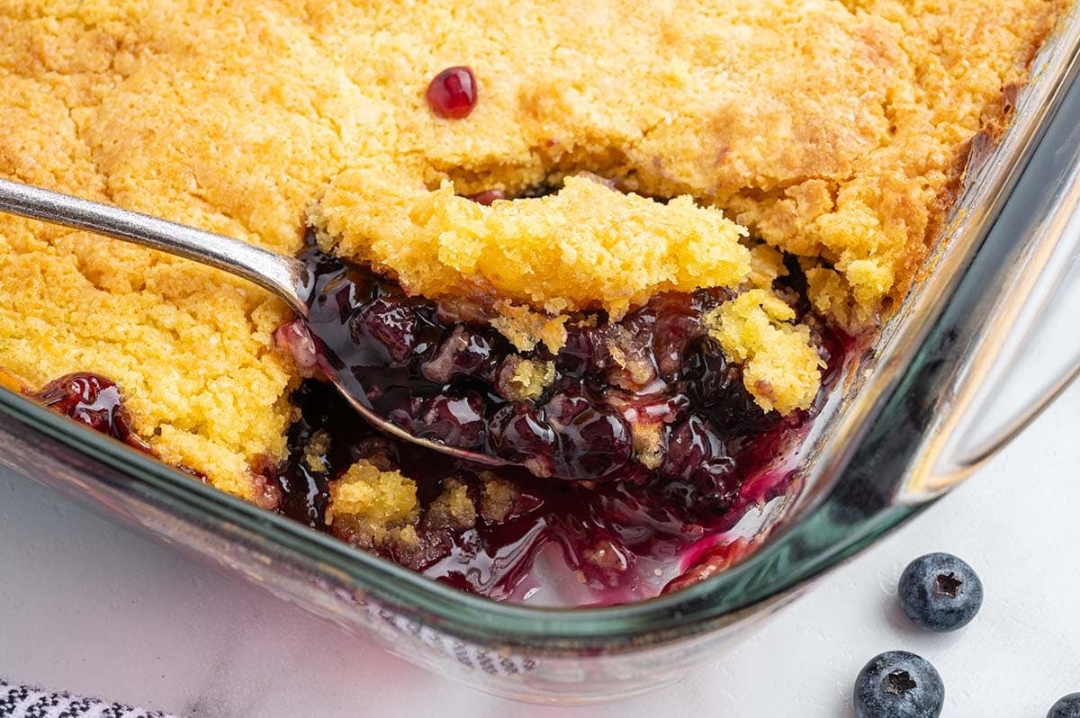 Blueberry dump cake being scooped from a glass baking dish showing the golden cake topping and blueberry filling.