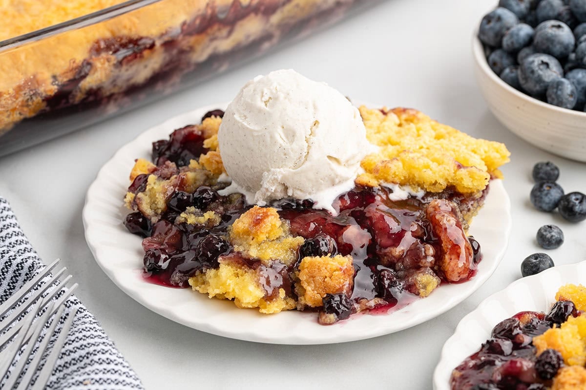 Serving of blueberry dump cake topped with a scoop of vanilla ice cream on a white plate with fresh blueberries nearby.
