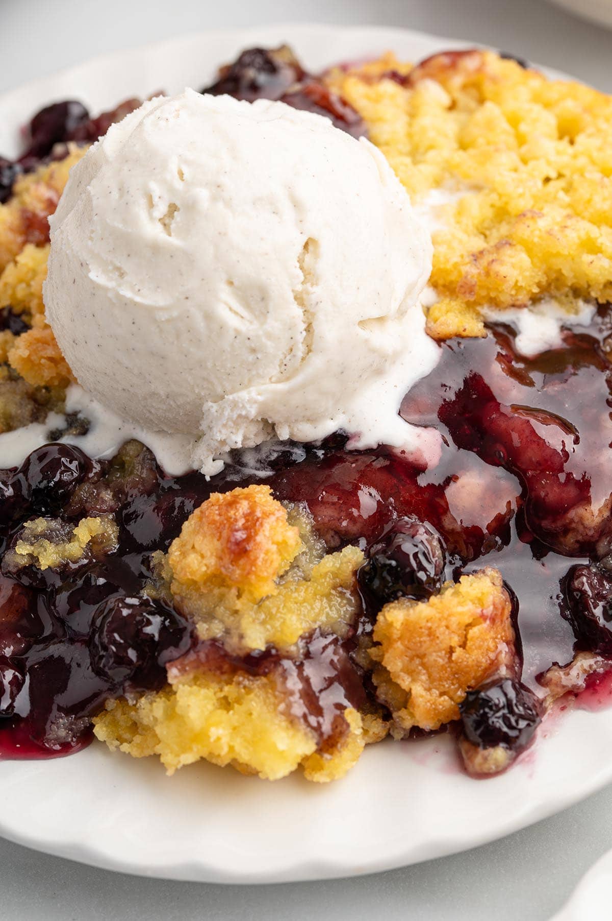 Close-up of blueberry dump cake with golden cake crumbles and a melting scoop of vanilla ice cream on a white plate.