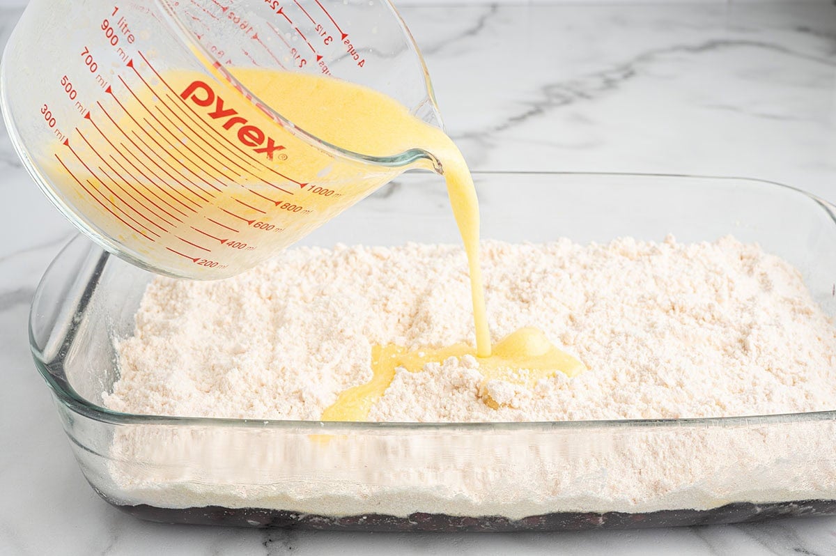 Melted butter being poured from a Pyrex measuring cup over dry cake mix in a glass baking dish.