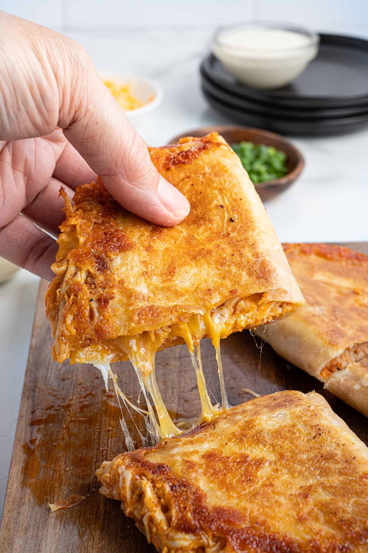 Hand lifting a crispy buffalo chicken quesadilla with melted cheese stretching from the toasted tortilla, served on a wooden board.