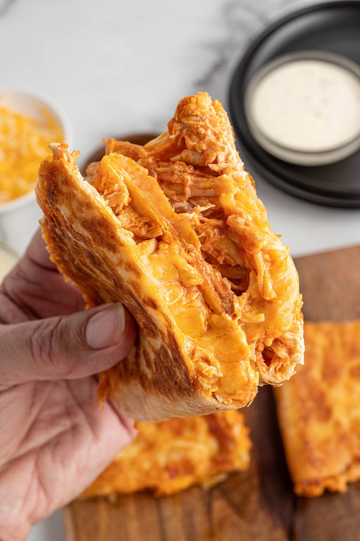 Hand holding a buffalo chicken quesadilla with melted cheese and shredded buffalo chicken inside a crispy toasted tortilla, with dipping sauce in the background.