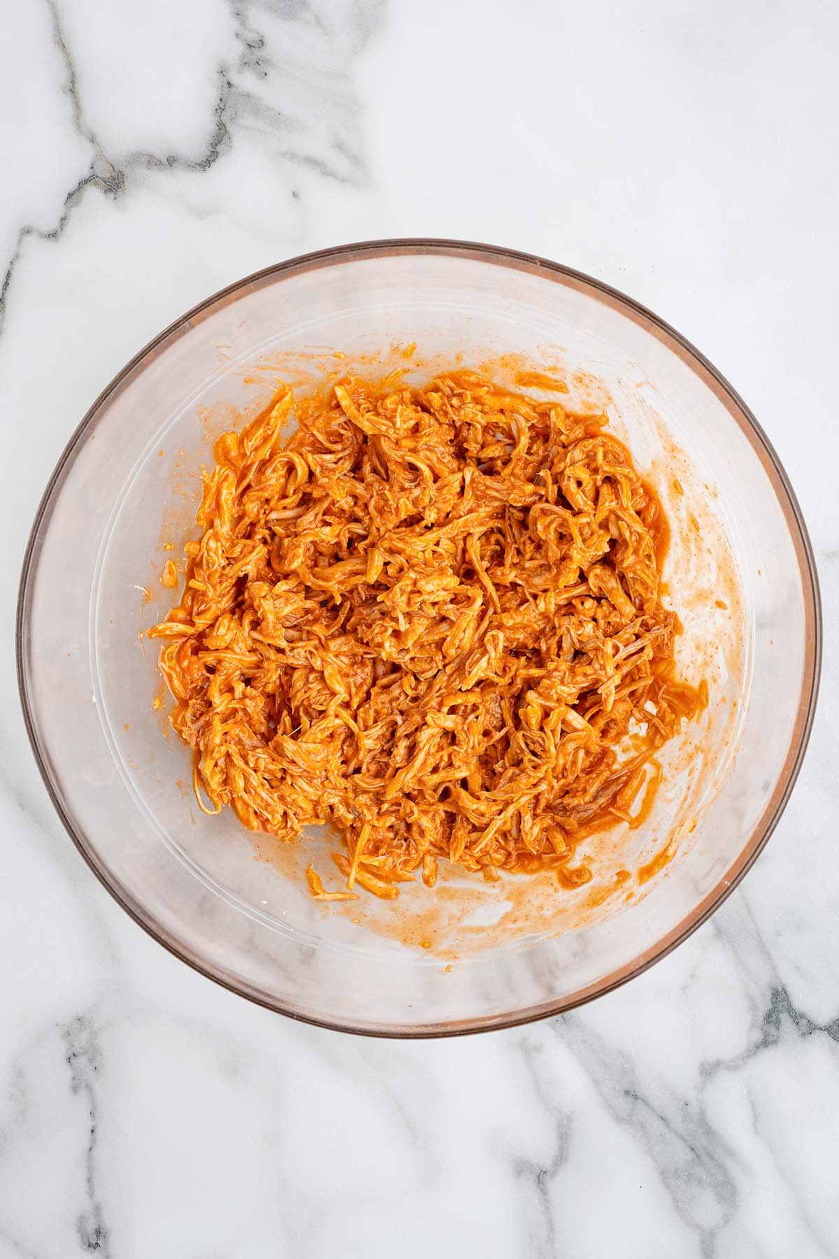 Overhead view of shredded chicken mixed with buffalo sauce in a glass bowl for making buffalo chicken quesadillas.