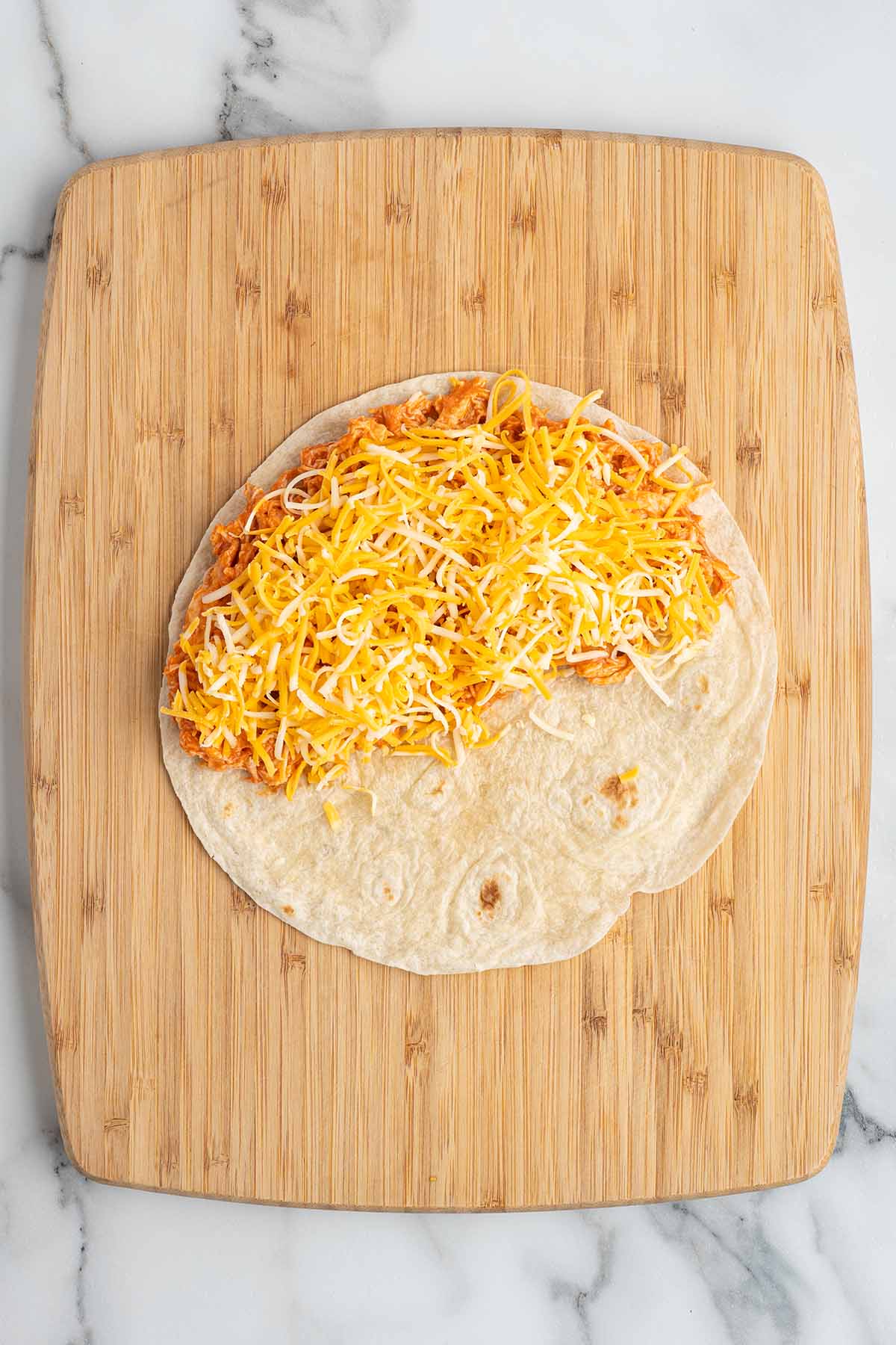 Overhead view of a tortilla filled with buffalo chicken and shredded cheese on a wooden cutting board before folding into a quesadilla.
