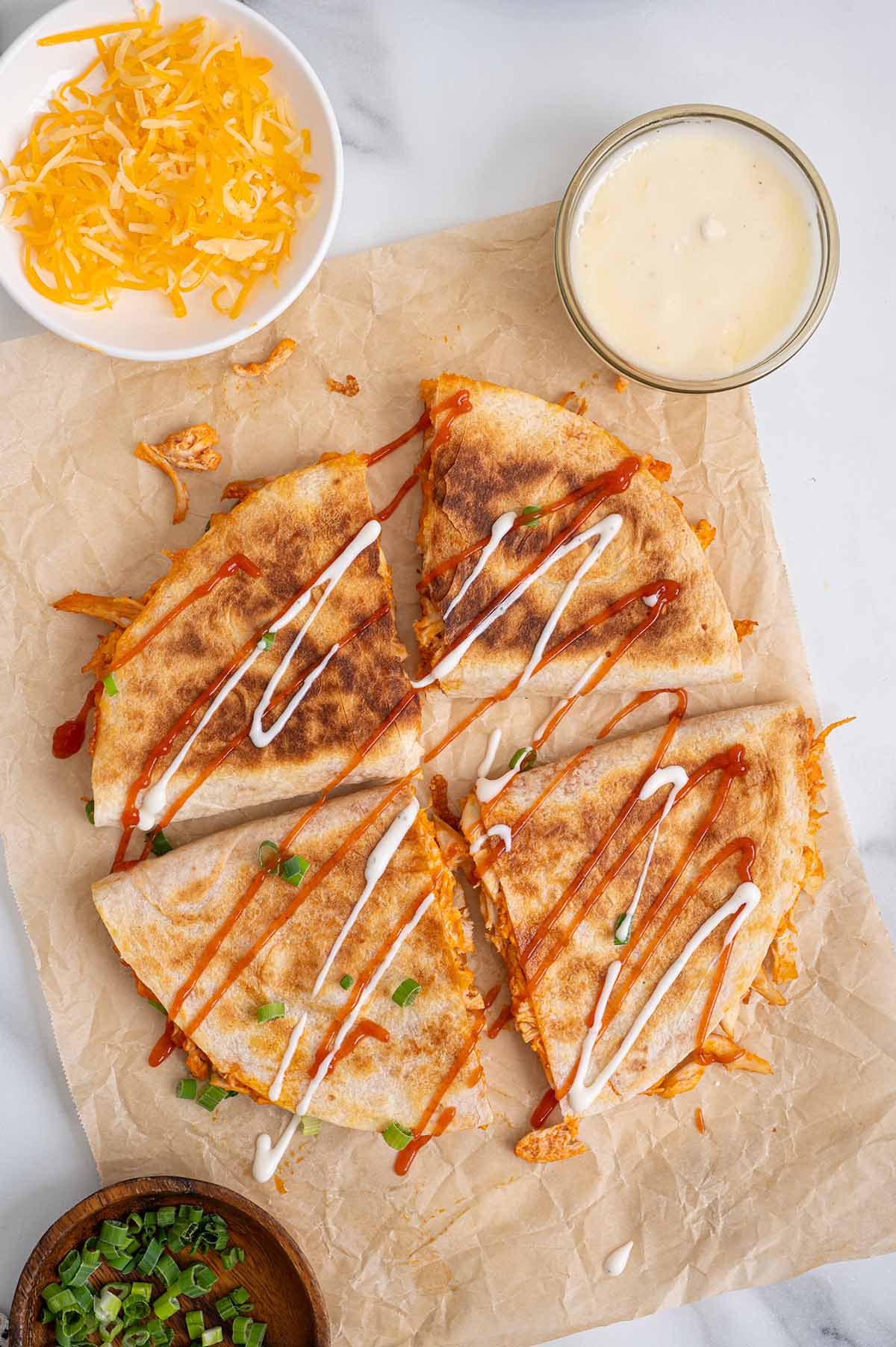 Overhead view of buffalo chicken quesadilla slices drizzled with buffalo sauce and ranch dressing, served with shredded cheese, green onions, and dipping sauce.