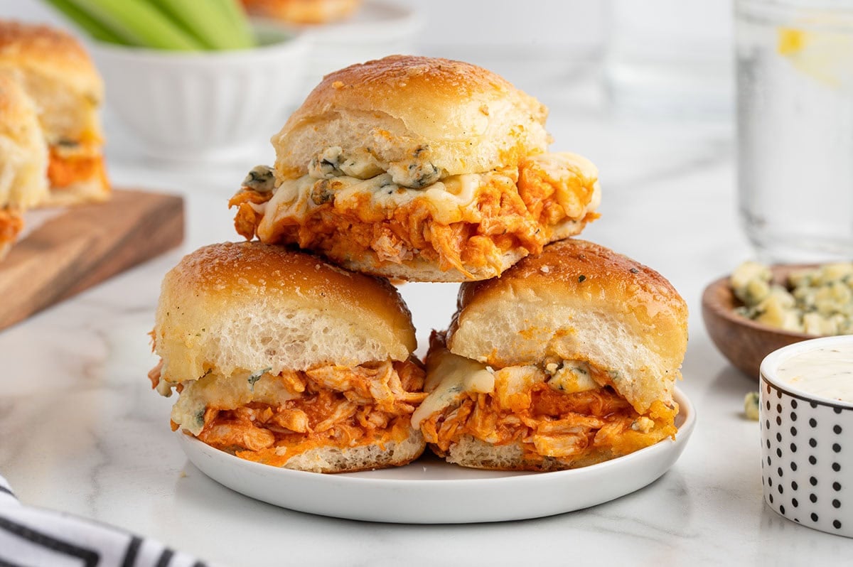 Three buffalo chicken sliders stacked on a plate with shredded buffalo chicken, melted cheese, and soft toasted slider buns, served with celery and dipping sauce.