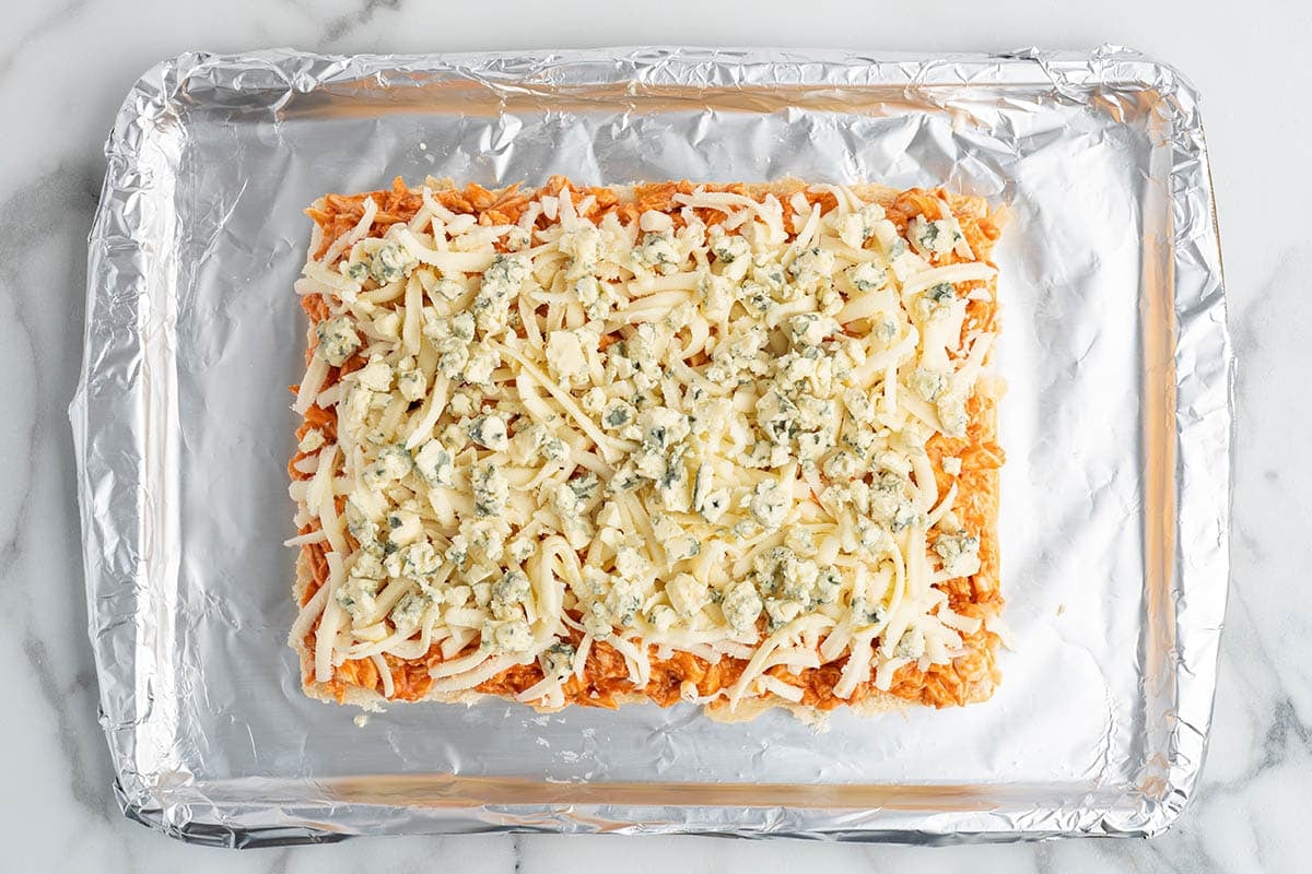 Overhead view of buffalo chicken sliders topped with shredded mozzarella and crumbled blue cheese on a foil-lined baking sheet before baking.