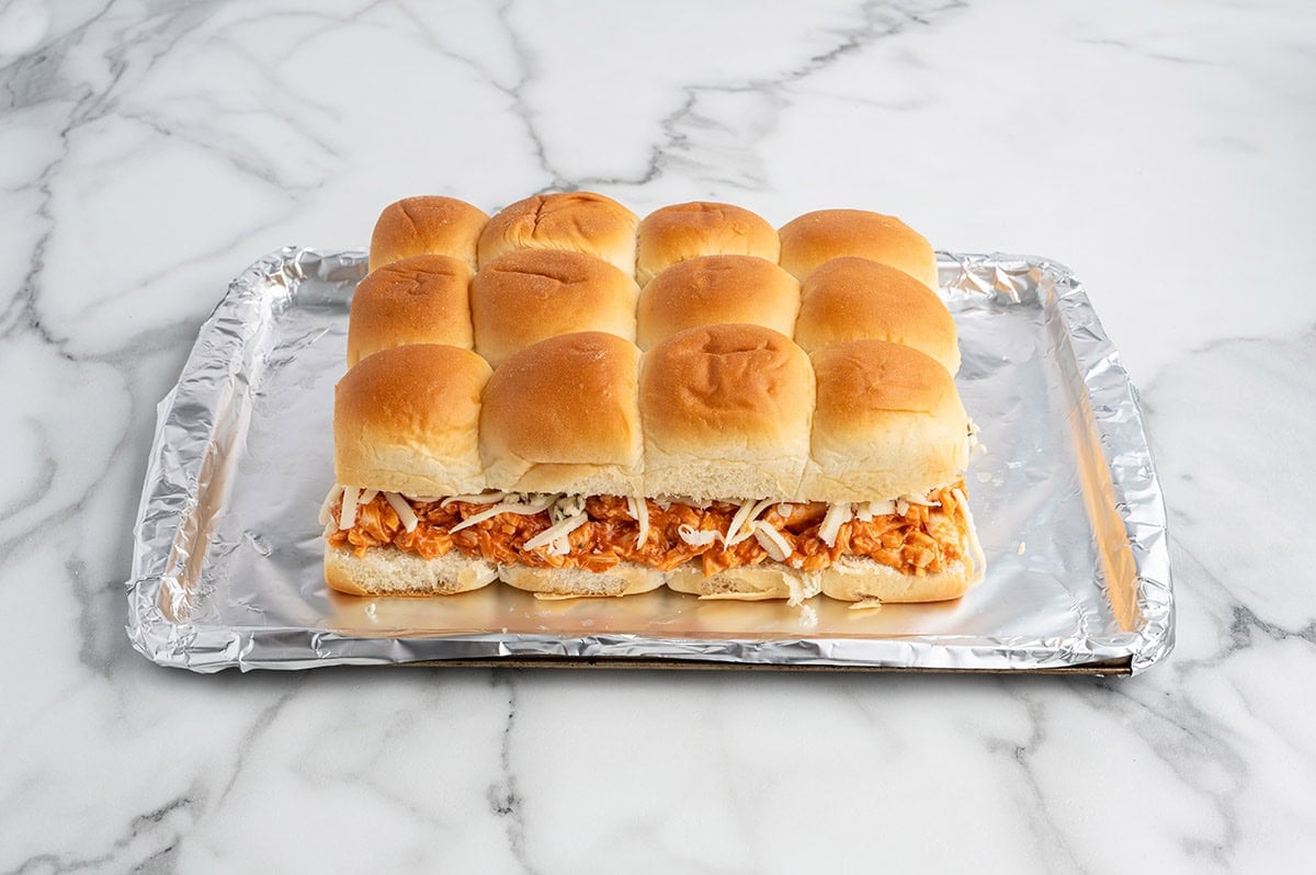 Unbaked buffalo chicken sliders assembled on a foil-lined baking sheet with shredded buffalo chicken, melted cheese, and soft slider buns on top.