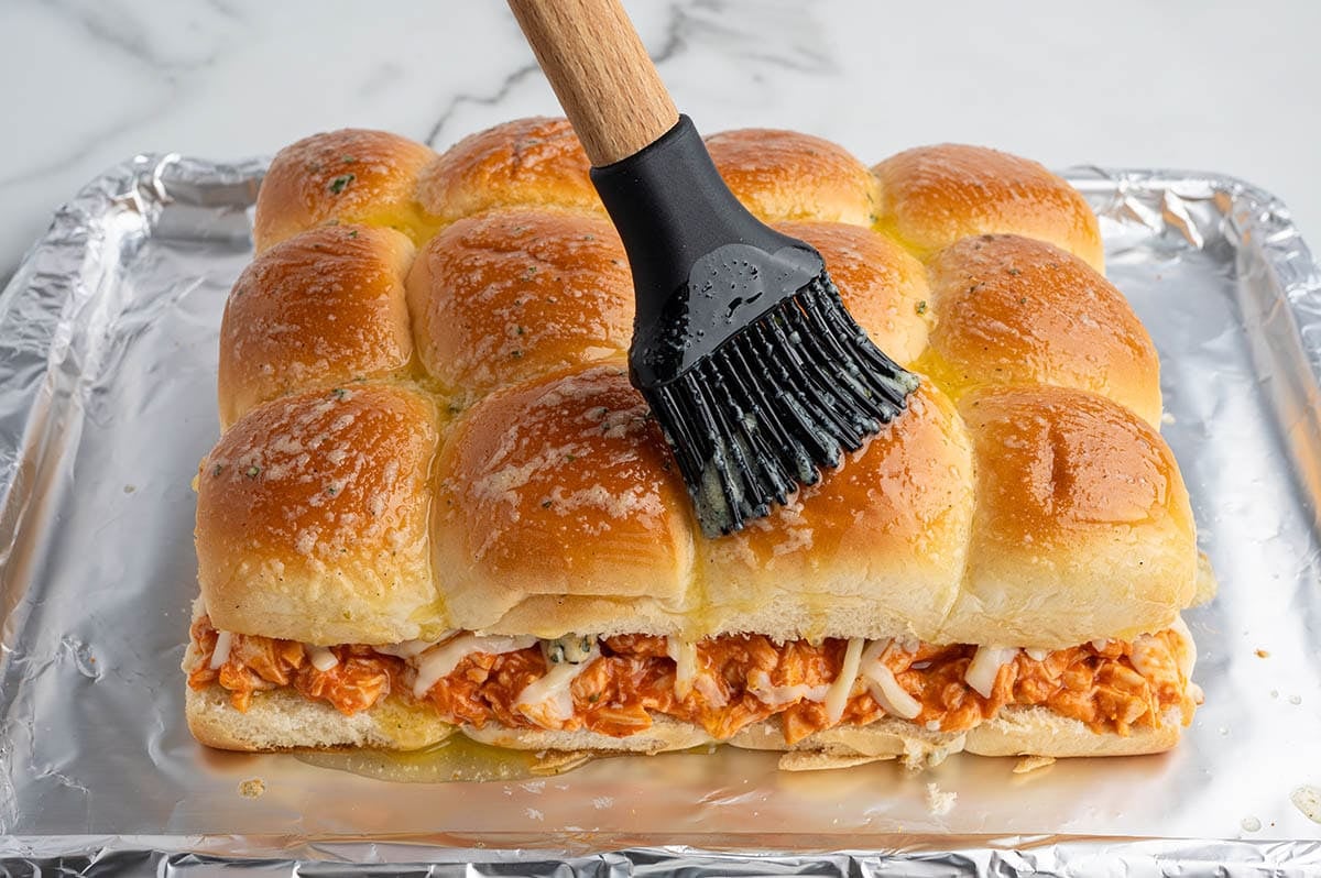 Buffalo chicken sliders on a foil-lined baking sheet being brushed with garlic butter before baking, filled with shredded buffalo chicken and melted cheese.