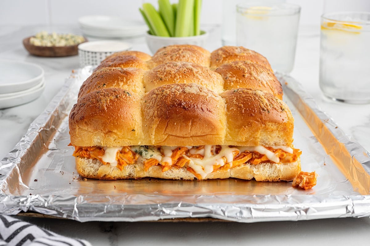 Baked buffalo chicken sliders on a foil-lined baking sheet with golden toasted buns, shredded buffalo chicken, and melted cheese, served with celery and dipping sauce.