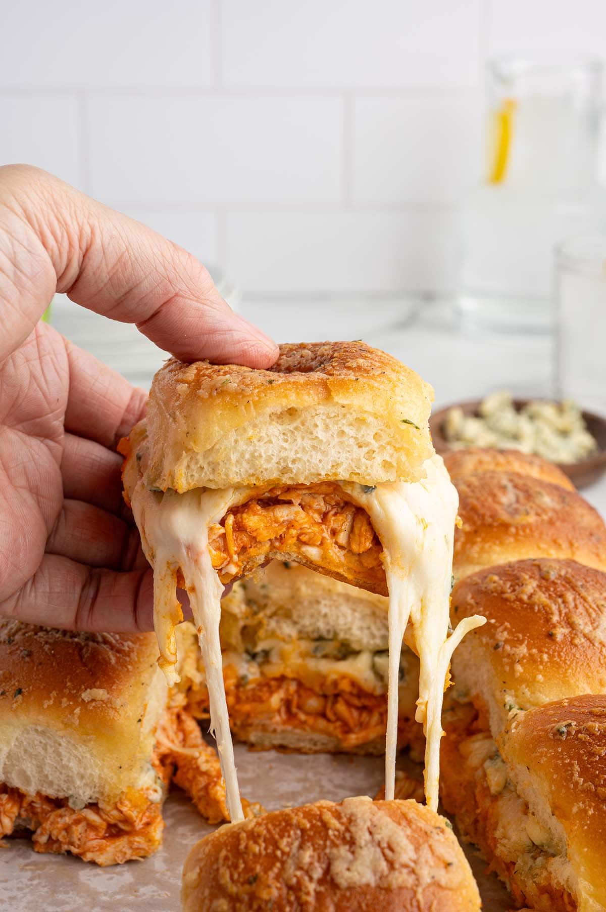 Hand holding a cheesy buffalo chicken slider with melted mozzarella stretching from the toasted bun, surrounded by baked buffalo chicken sliders.