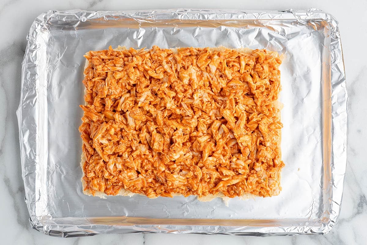 Overhead view of shredded buffalo chicken layered onto slider buns on a foil-lined baking sheet for buffalo chicken sliders.