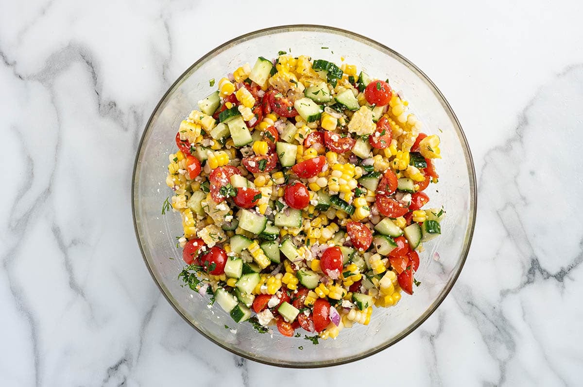Overhead view of corn salad with cherry tomatoes, cucumber, red onion, and feta in a glass bowl.