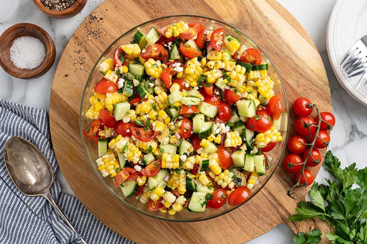 Overhead view of fresh corn salad with cherry tomatoes, cucumber, and feta in a glass bowl on a wooden serving board.