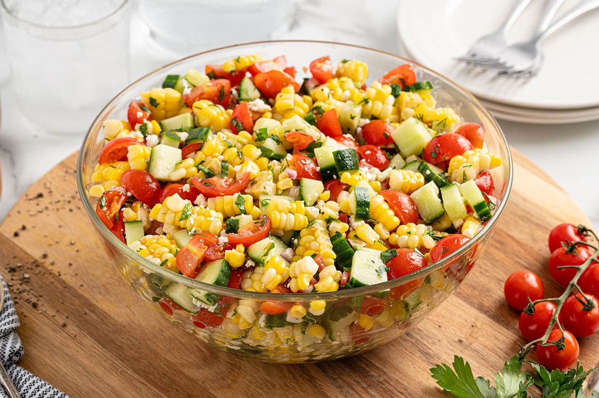 Corn salad with cherry tomatoes, cucumber, feta and fresh herbs in a glass bowl on a wooden cutting board.