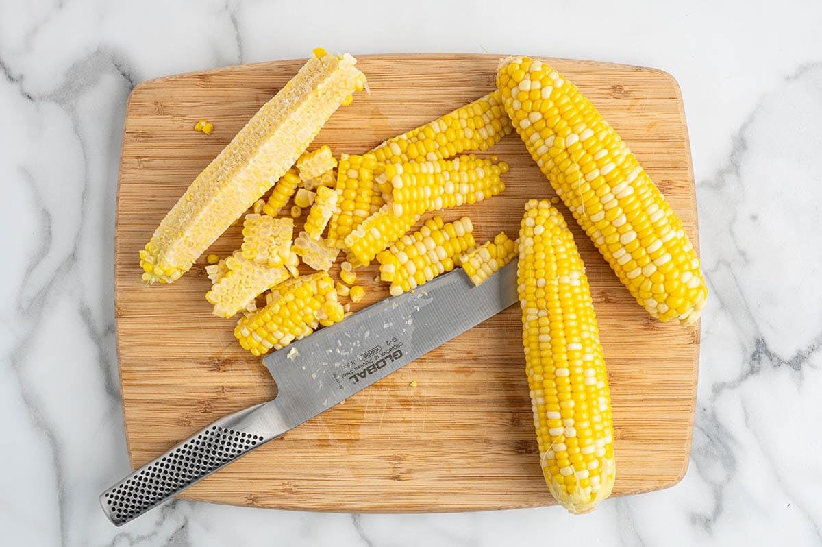 Fresh corn being cut off the cob on a wooden cutting board with a chef's knife.