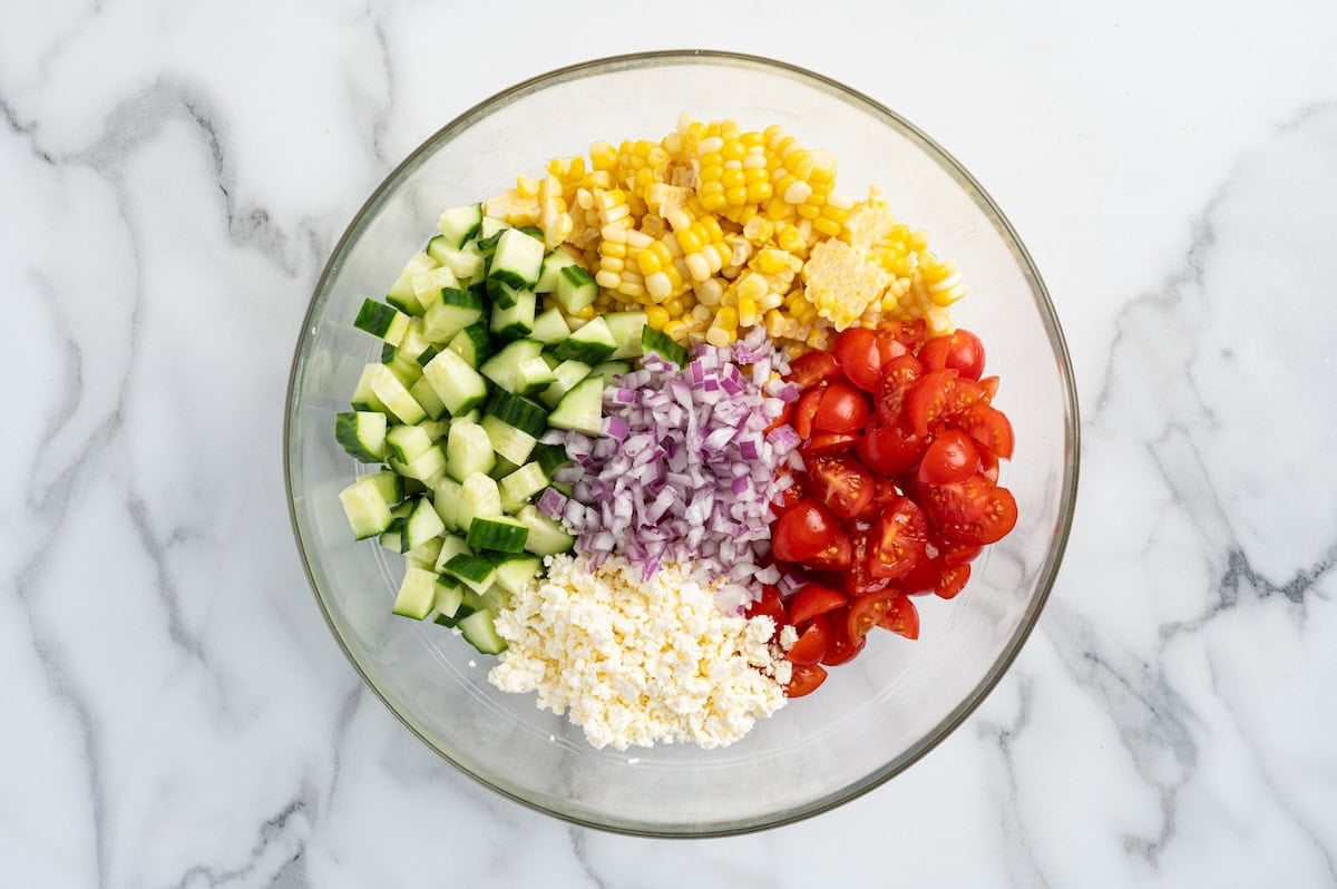 Corn salad ingredients arranged in a glass bowl — fresh corn, cucumber, cherry tomatoes, red onion, and crumbled feta before mixing.