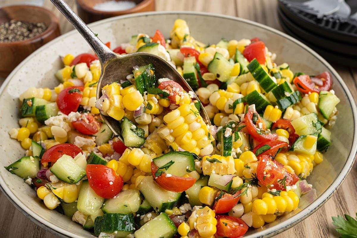 Close-up of corn salad with cherry tomatoes, cucumber, and feta being served with a spoon.