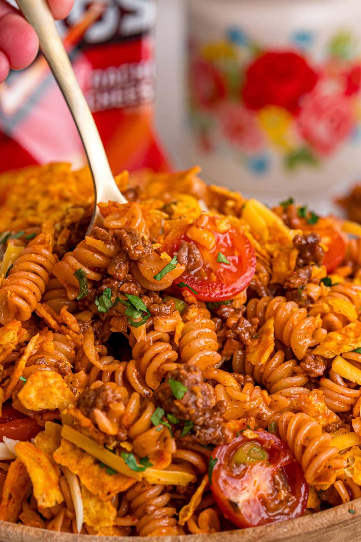 Close-up of Doritos pasta salad being served with a spoon, showing rotini, ground beef, cherry tomatoes, and crushed Doritos.
