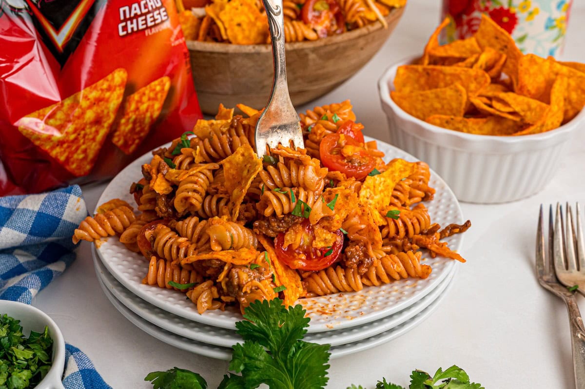 A serving of Doritos pasta salad on a white plate with a fork, with the serving bowl and a bag of Doritos in the background.