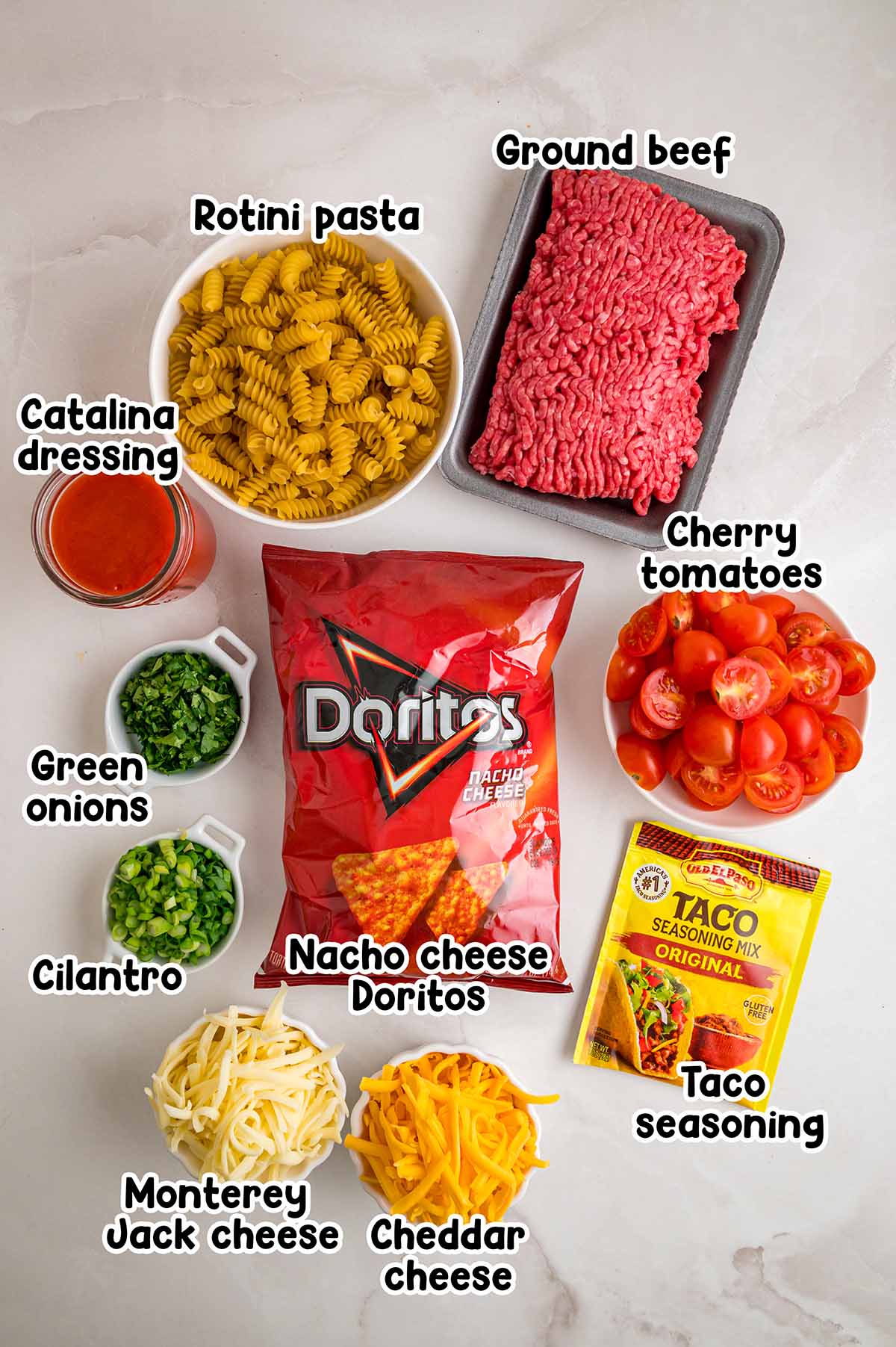 Labeled ingredients for Doritos pasta salad including rotini, ground beef, nacho cheese Doritos, cherry tomatoes, Catalina dressing, taco seasoning, cheddar, Monterey Jack, green onions, and cilantro.