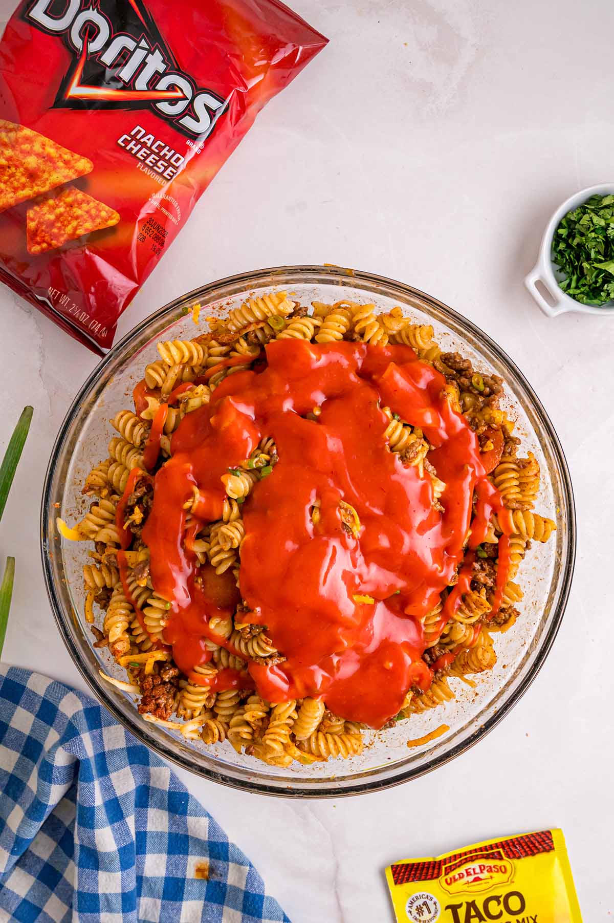 Catalina dressing being poured over rotini and ground beef in a glass bowl for Doritos pasta salad.