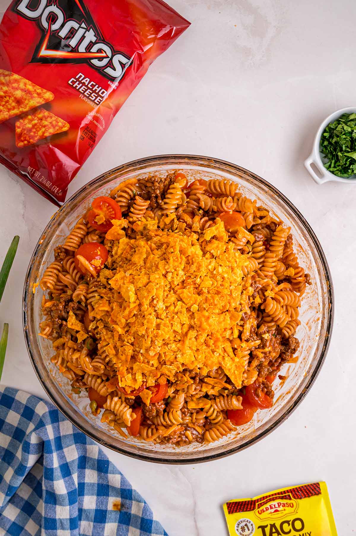 Overhead view of Doritos pasta salad topped with crushed nacho cheese Doritos in a glass bowl with a bag of Doritos and taco seasoning packet nearby.