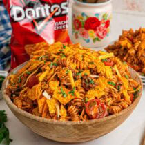 Wooden bowl piled high with Doritos pasta salad with rotini, ground beef, cherry tomatoes, and Dorito chips, with a bag of nacho cheese Doritos in the background.