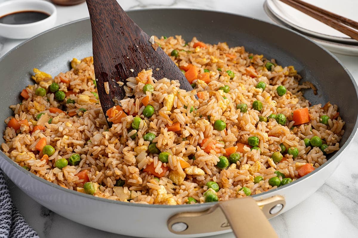 Skillet filled with homemade vegetable fried rice made with carrots, peas, scrambled eggs, onions, and soy sauce, being stirred with a wooden spatula.