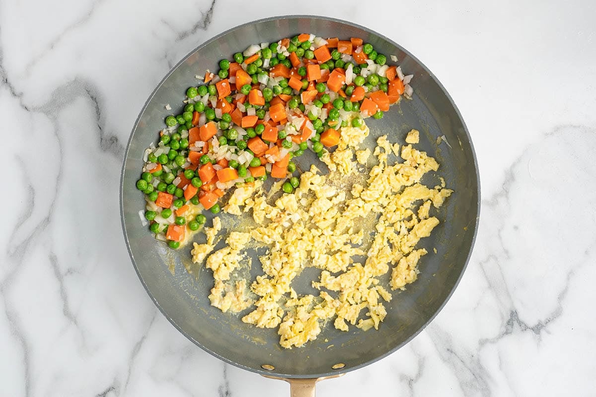 Overhead view of scrambled eggs, peas, carrots, and onions cooking in a skillet as the base for homemade fried rice.