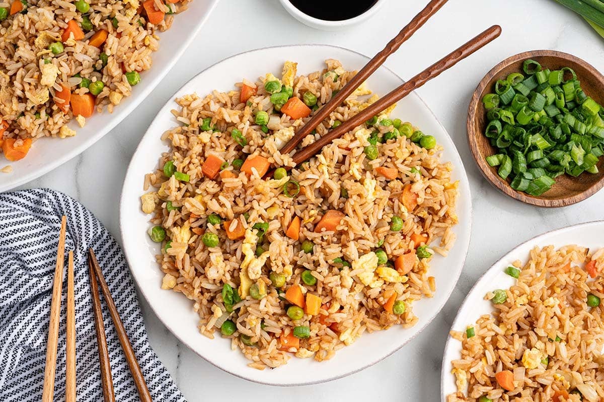 Overhead view of homemade fried rice with carrots, peas, scrambled eggs, and green onions served on plates with chopsticks and soy sauce on the side.