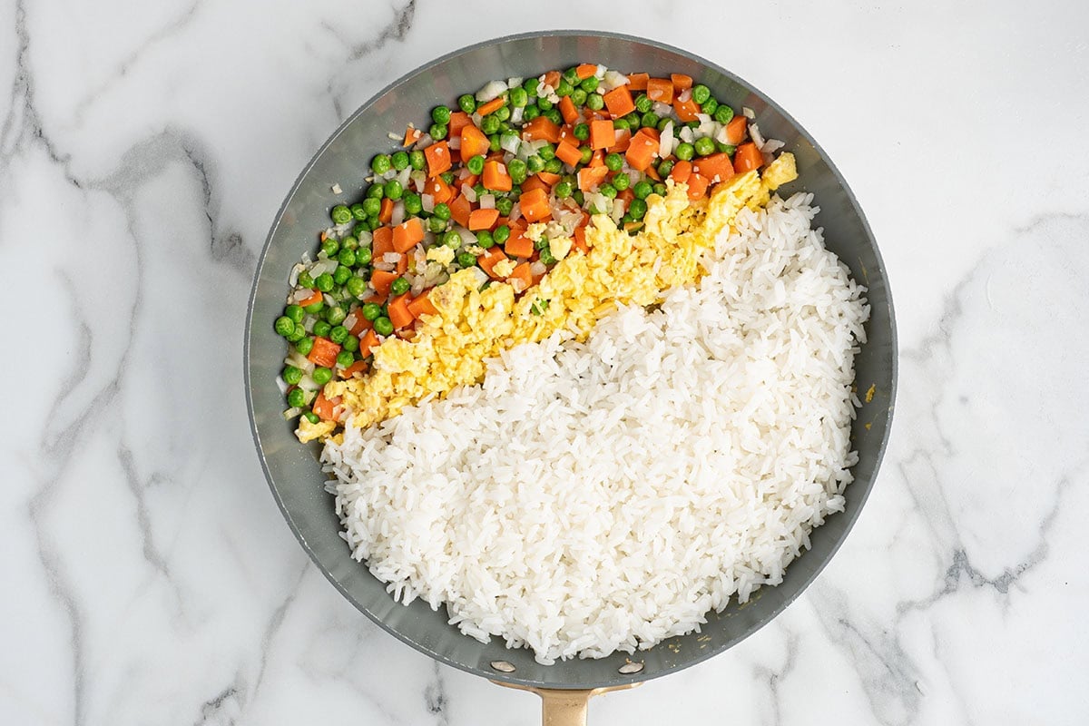 Overhead view of cooked white rice, scrambled eggs, peas, carrots, and onions in a skillet before being mixed into homemade fried rice.
